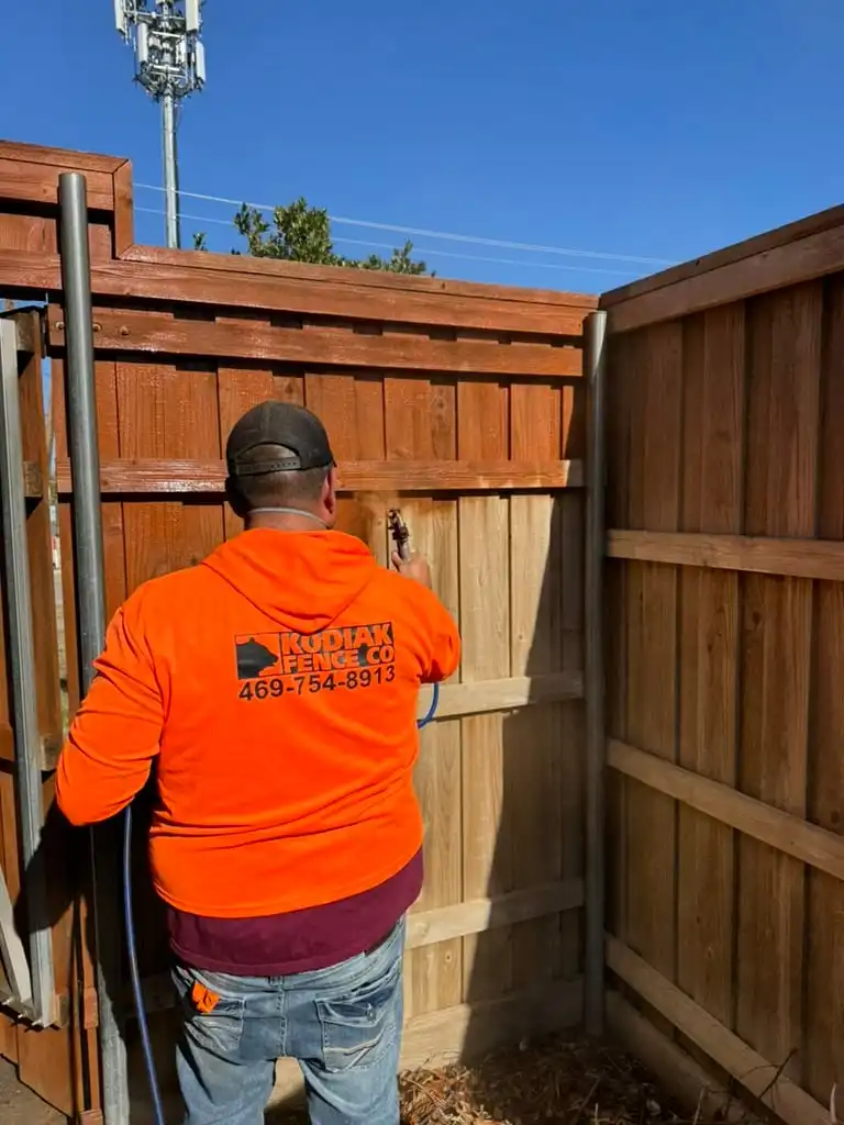 Kodiak crew member spraying stain onto a cedar fence, half the boards already colored