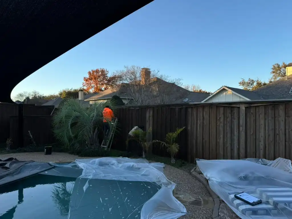 Crew staining a tall board-on-board fence with the pool and patio furniture fully covered